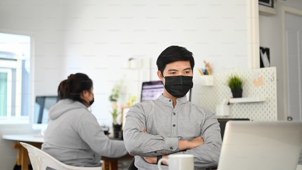 Ein junger Mann mit Gesichtsmaske hält die Arme verschränkt und schaut auf den Laptop-Bildschirm, während er im Büro sitzt.