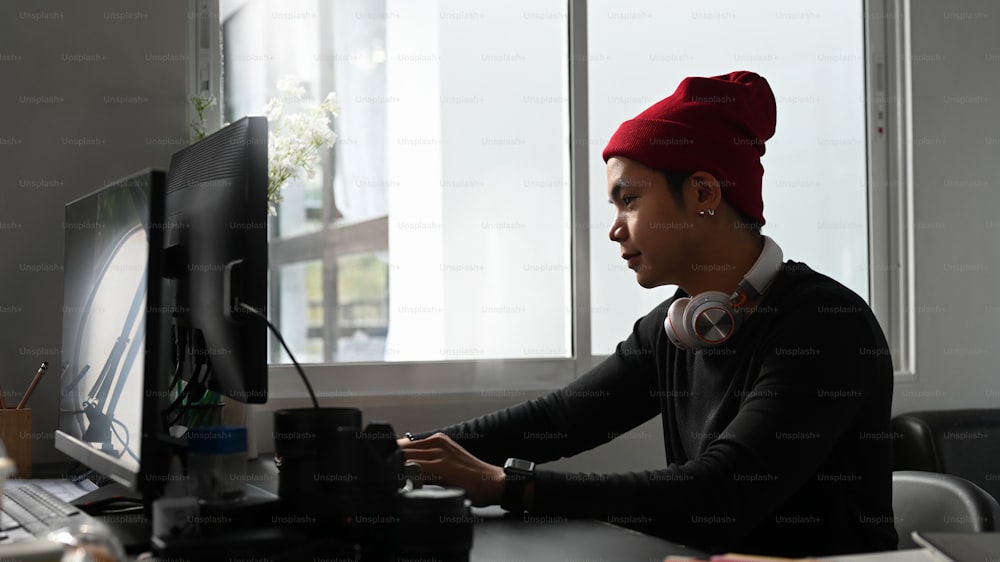 Seitenansicht eines jungen Fotografen mit roter Wollmütze, der in seinem Studio an einem neuen Projekt am Computer arbeitet.