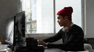 Side view of young photographer in red wool hat working with new project on computer at his studio.