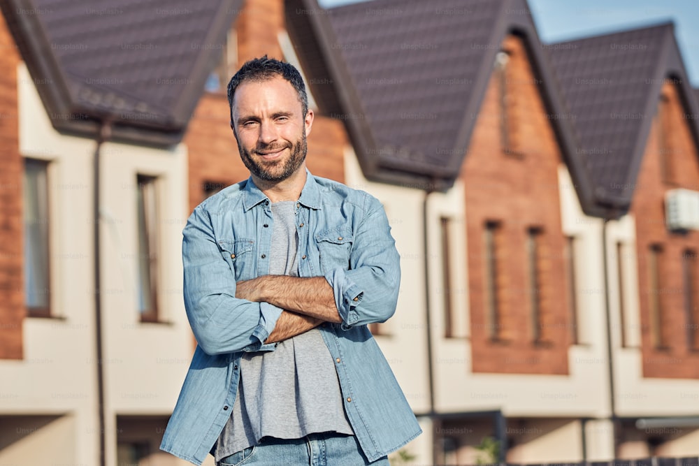 Contented brunette man standing cross-armed in front of a lovely house