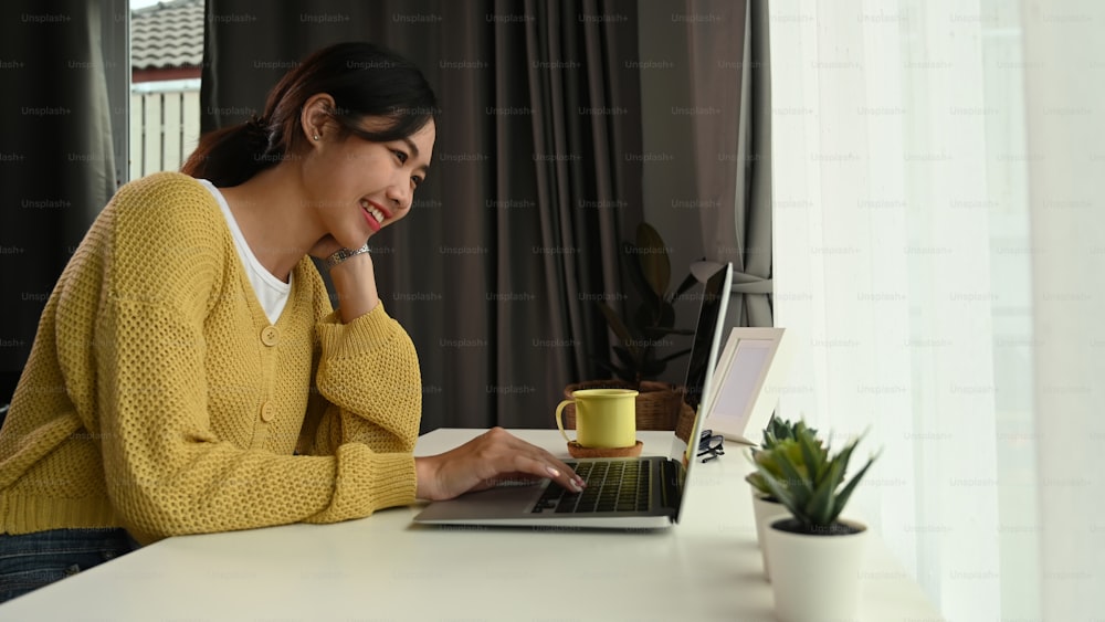 Side view of happy young woman looking on screen of laptop while sitting at home office.