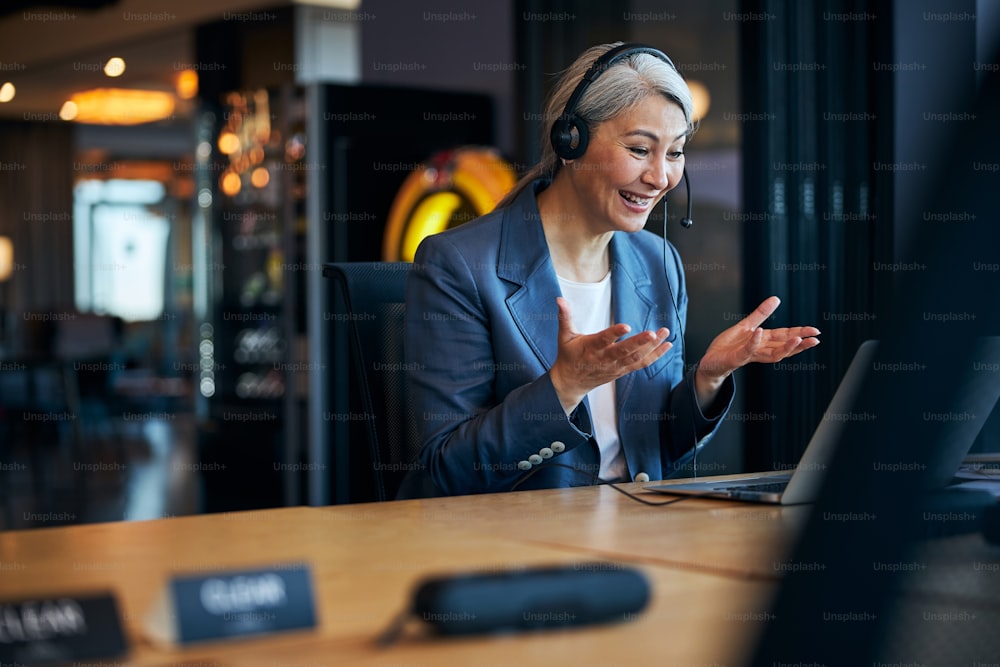 Cheerful businesswoman talking with business partner through video call and smiling while sitting at the table with laptop