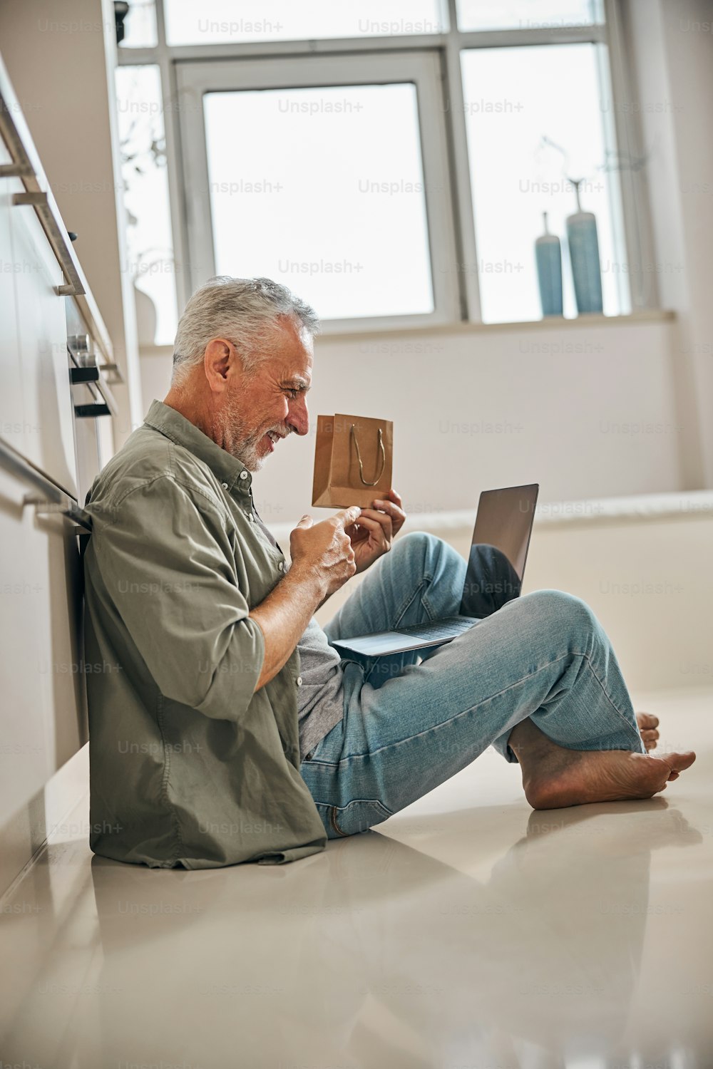 Anciano optimista que sostiene una pequeña bolsa de regalo frente a la pantalla de una computadora portátil y gesticula activamente