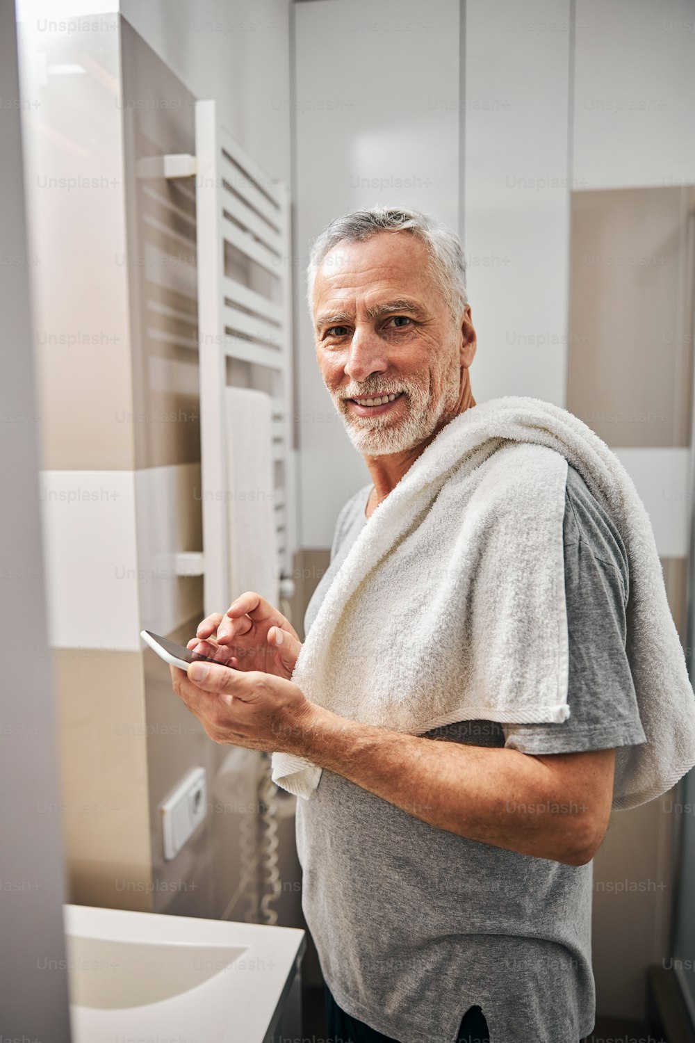 Waist-up photo of a cheerful aged man with a towel standing in the bathroom and holding a phone