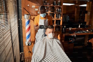 Jovem bonito usando máscara facial protetora enquanto corta o cabelo em barbearia durante a pandemia