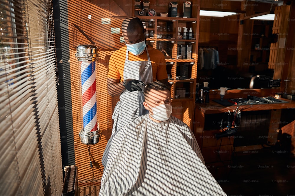 Hübscher junger Mann trägt eine schützende Gesichtsmaske beim Haarschnitt im Friseursalon während der Pandemie