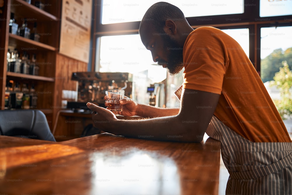 Bearded Afro American man with glass of alcoholic drink in his hand standing at the bar counter