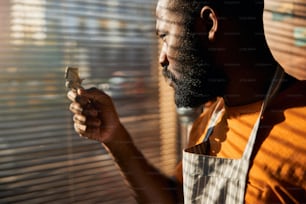 Close up of handsome male barber looking at hair clipper with serious expression while standing by the window with blinds