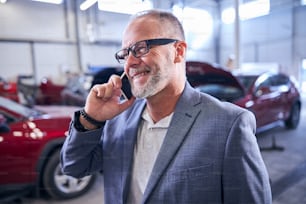 Handsome gentleman in glasses talking on cellphone and smiling while standing in vehicle repair shop
