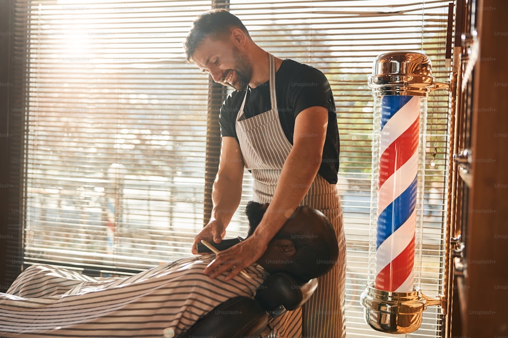 Bearded Afro American man sitting in salon chair while joyful male barber removing stubble from his neck with electric hair trimmer