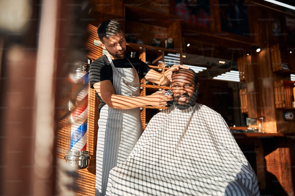 Handsome Afro American man sitting in barber chair and smiling while hairdresser removing stubble from his cheek with professional shaving machine