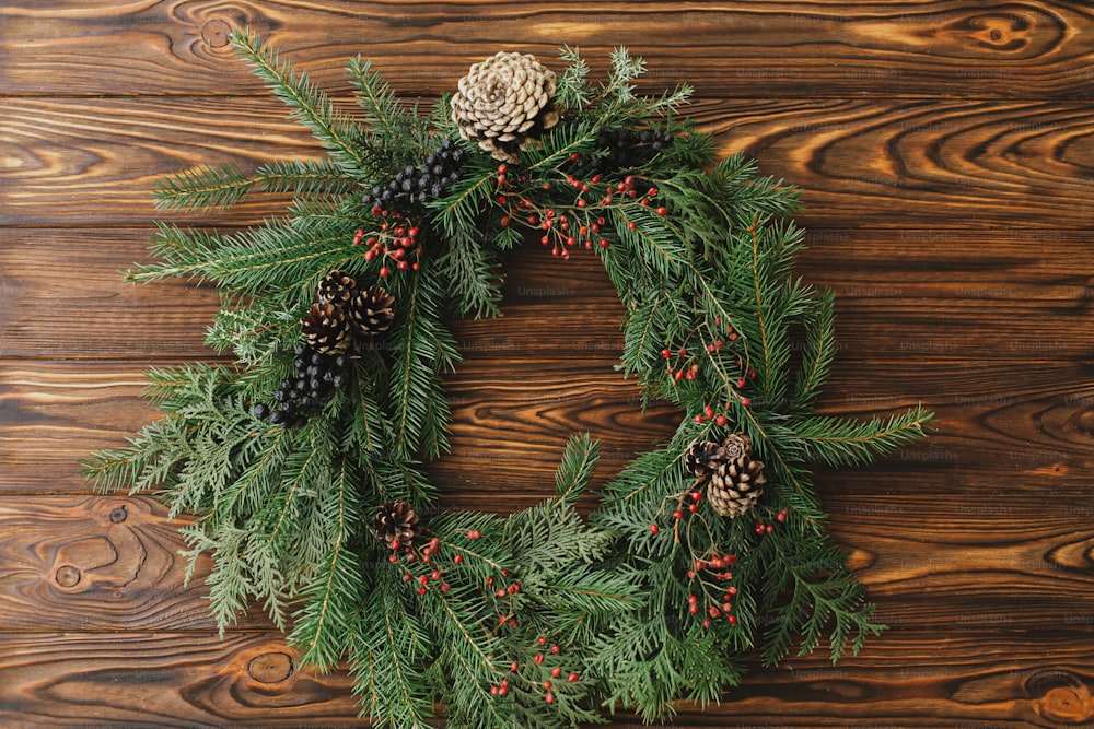 Rustikaler Weihnachtskranz auf Holztisch flach gelegt. Moderner Weihnachtskranz mit roten Beeren, grünen Zweigen, Tannenzapfen auf rustikalem Hintergrund. Frohe Weihnachten und schöne Feiertage