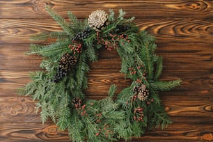 Rustic christmas wreath on wooden table flat lay. Modern christmas wreath with red berries, green branches, pine cones on rustic background. Merry Christmas and happy holidays