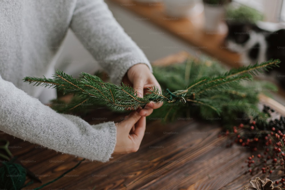 소박한 크리스마스 화환 만들기, 계절 겨울 강림. 녹색 전나무 가지를 들고 나무 테이블을 배경으로 크리스마스 화환을 만드는 여성 손을 닫습니다. 수제 축제 장식