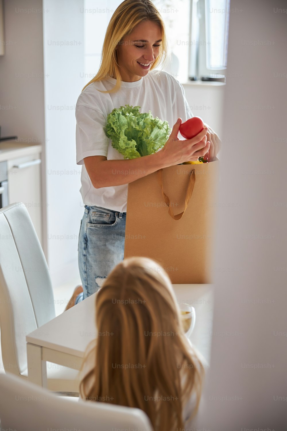 Mujer hermosa caucásica adulta de pie cerca de la mesa blanca de la cocina con una bolsa de compras de papel mientras sostiene verduras en las manos