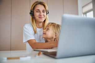 Waist up portrait of blonde Caucasian woman in headphones with little girl sitting near her working from home office at the quarantine time