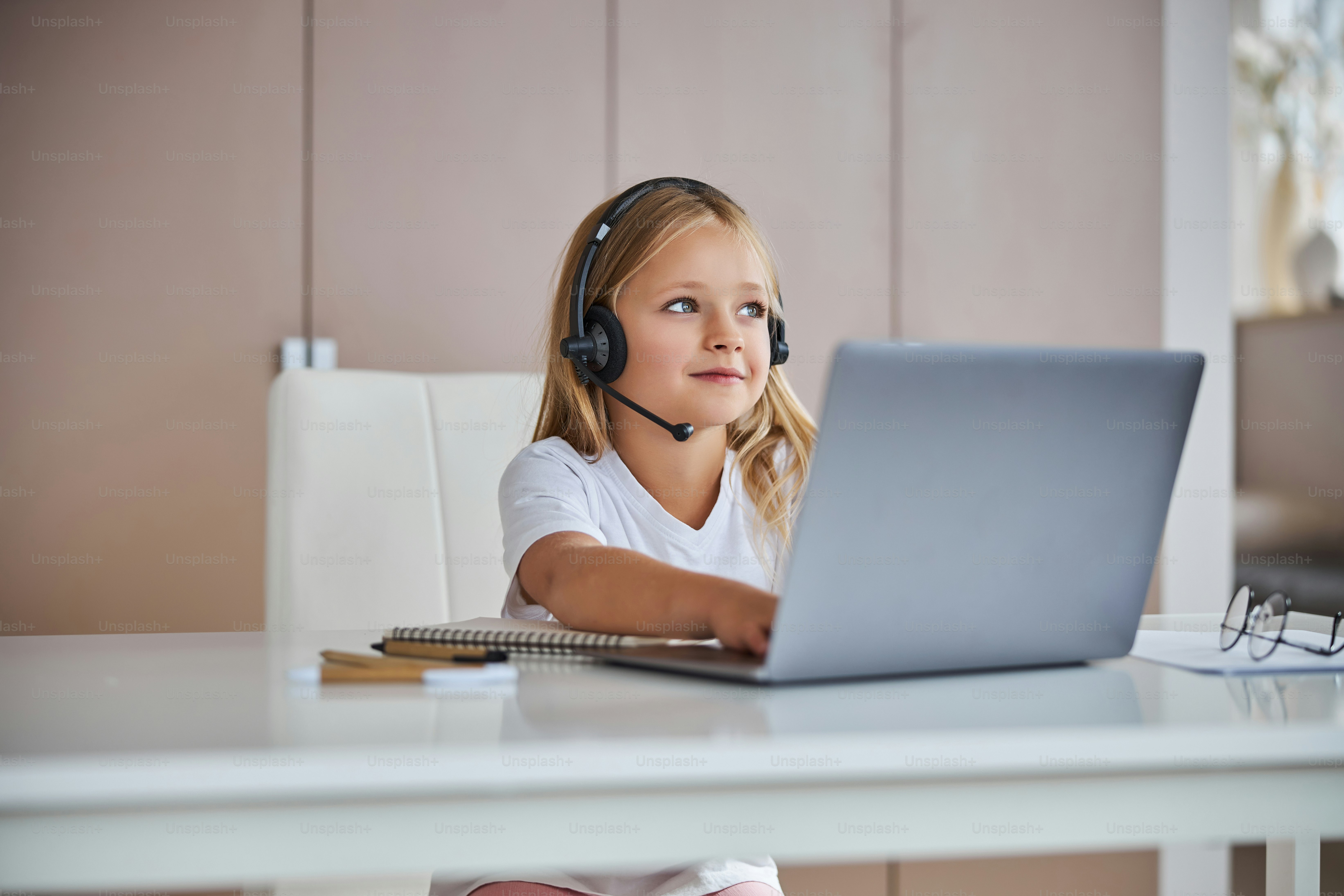 Little cute school girl spending time at the laptop while looking away and listening something in headphones at the work desk in her room