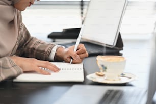 Cropped sot of a young Arabic business woman wearing hijab is taking notes in notebook at her workspace.