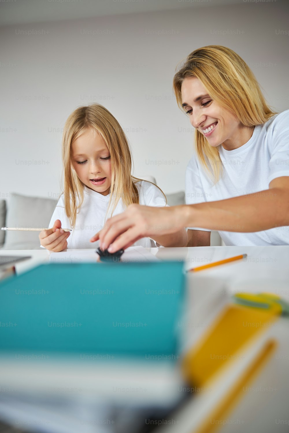 Side view portrait of attractive smiling Caucasian woman in casual clothes enjoying time with little girl while drawing something in exercises book in apartment