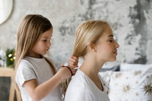 Concept of tender moments. Profile portrait of focused girl braids hair and making hairstyle for her dreamy mother. Charming daughter, spending free time together with mom and playing at home