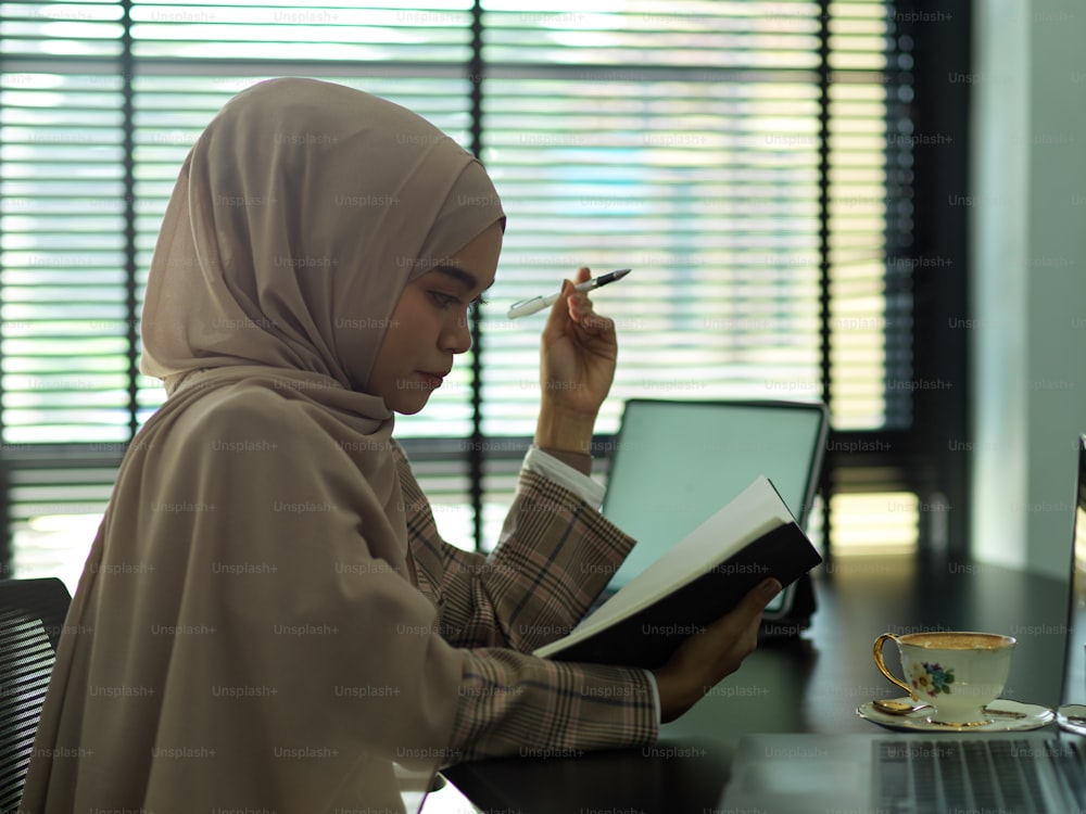 Vista laterale della donna d'affari musulmana asiatica che legge i documenti mentre è seduta nella stanza dell'ufficio