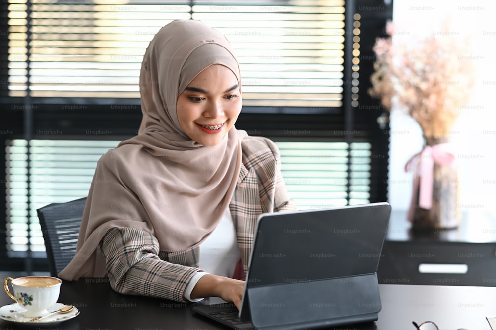 ヒジャブをかぶった若いアラブ人女性起業家がオフィスに座り、タブレットを使ってオンラインで仕事をしています。