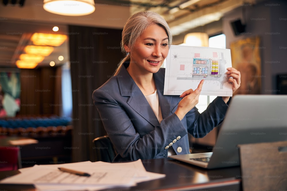 Beautiful businesswoman pointing at architecture building plan on sheet of paper while talking with business partner online