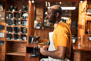 Bearded male barber in apron looking away and smiling while holding portable hair clipper