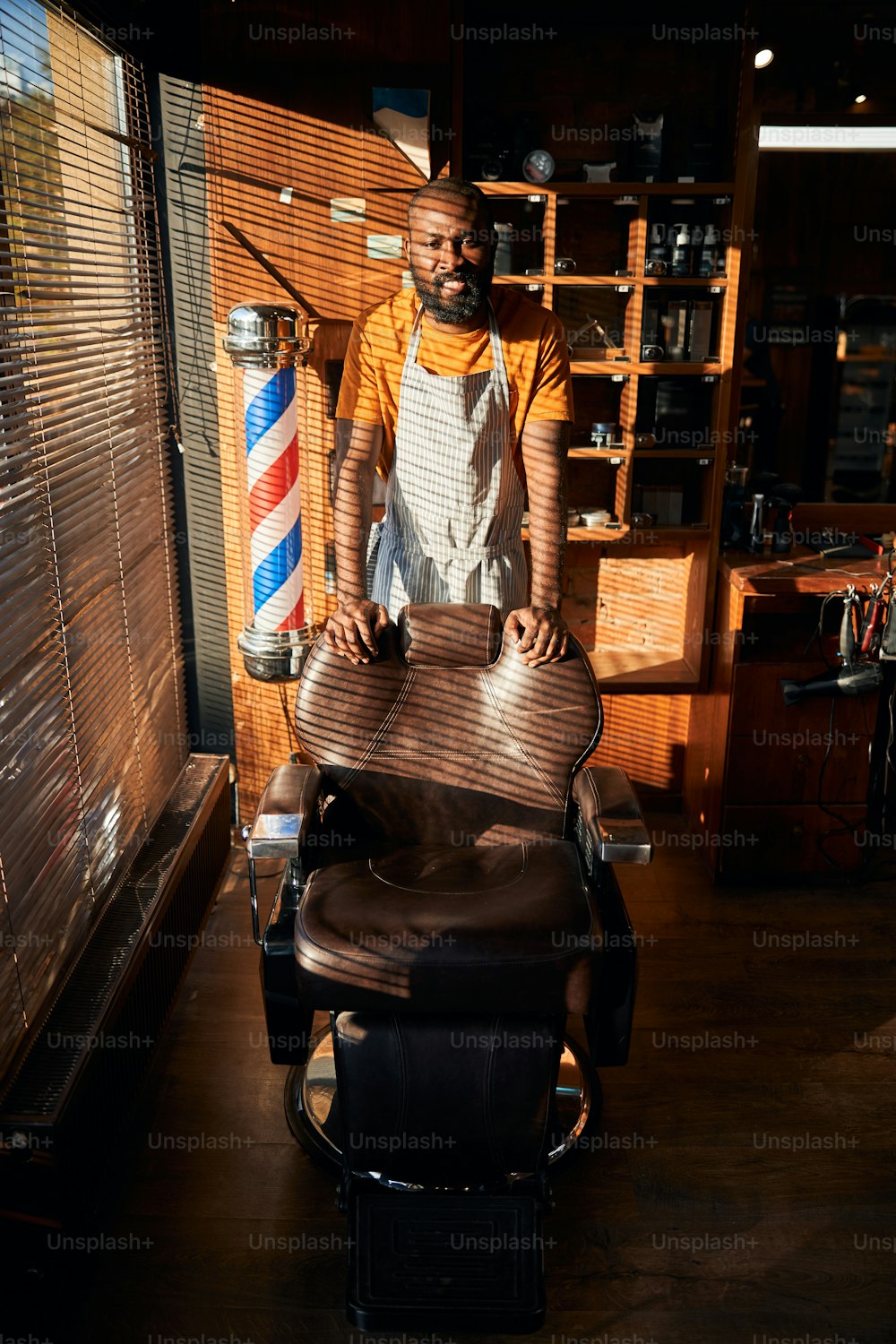 Homem afro-americano bonito em avental olhando para a câmera e sorrindo enquanto coloca as mãos na cadeira de barbeiro