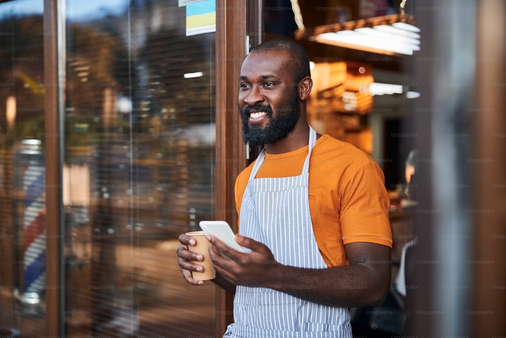 Handsome Afro American man in apron looking away and smiling while holding cup of coffee and cellphone