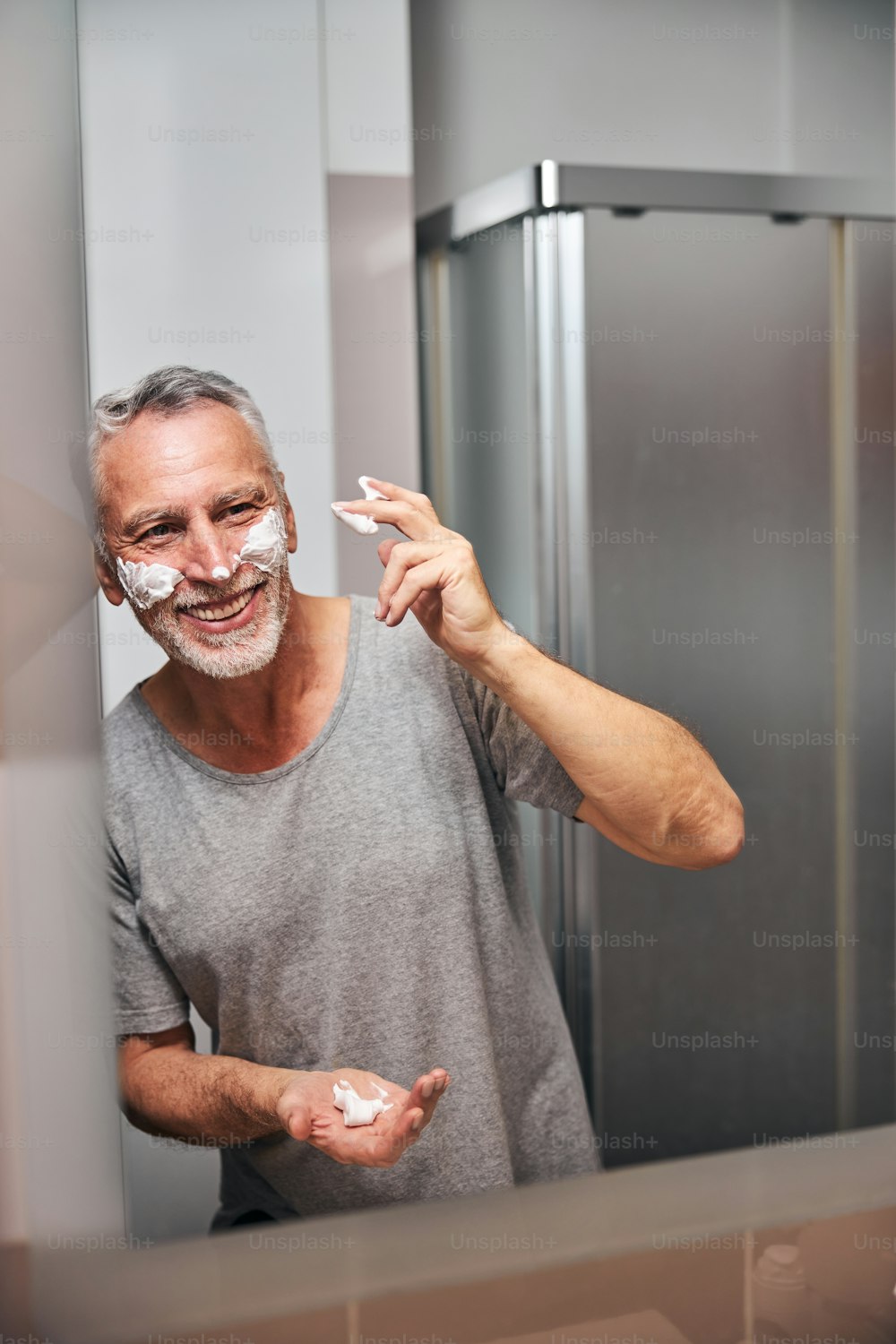 Optimistic aged man laughing while looking in the mirror and putting shaving foam on his face