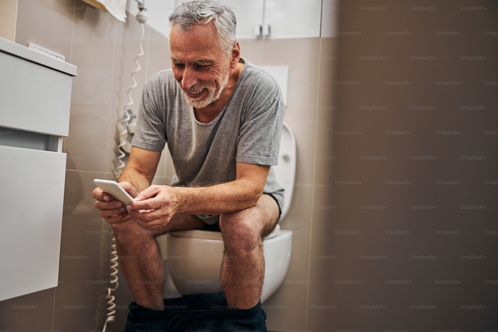 Grey-haired aging man laughing while sitting on the toilet seat and looking at his smartphone