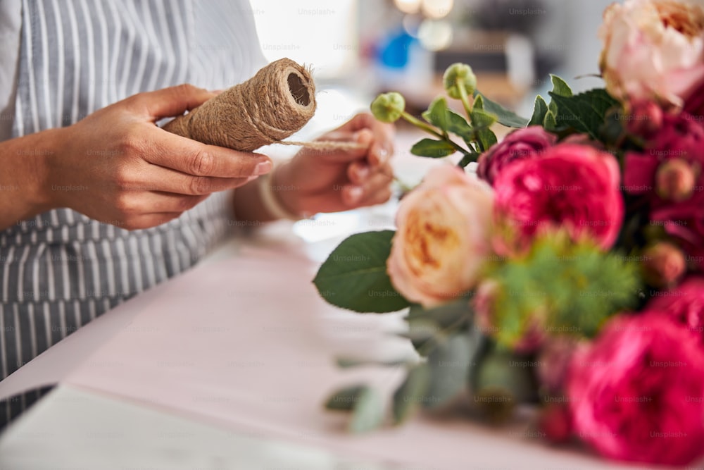Cropped photo of florist hands holding a spool of thread near a bunch of pretty flowers on a table