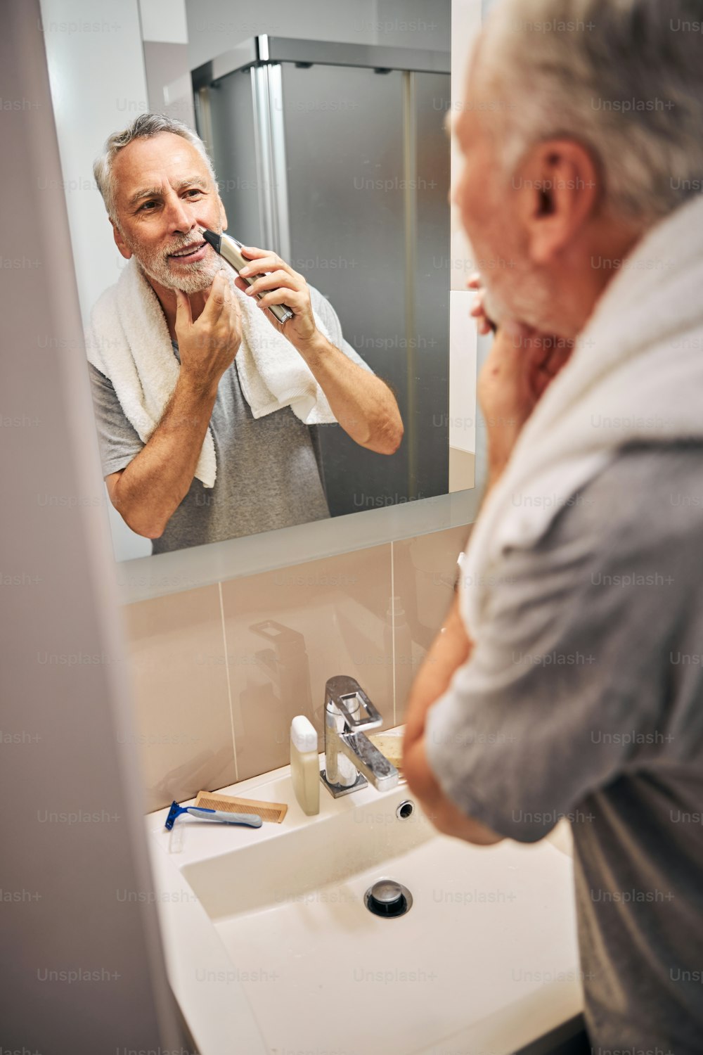 Positive-looking aged man looking in the mirror while trimming his nose hair with an elcetric device