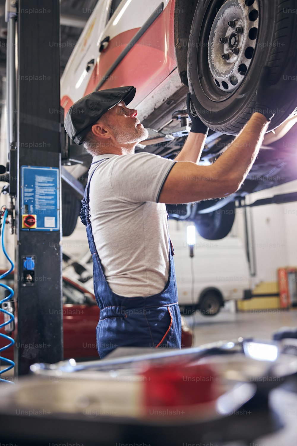 Handsome male technician in cap working with lifted car in automobile repair garage