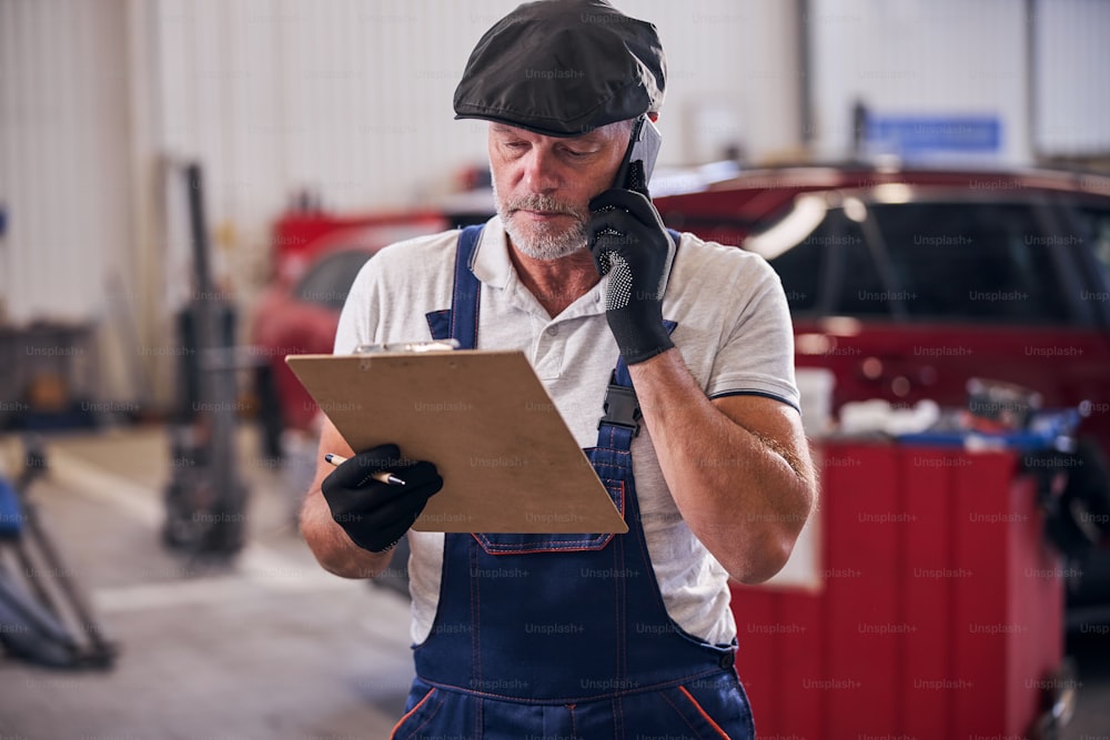 Trabajador masculino barbudo revisando notas mientras tiene una conversación telefónica en una estación de servicio de reparación de automóviles