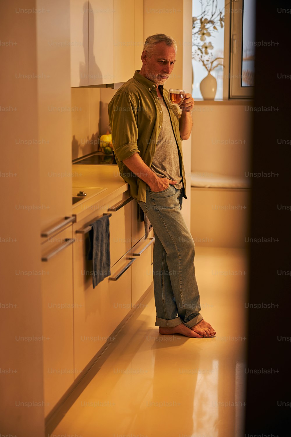 Relaxed elderly man having one hand in pocket and holding cup of tea while leaning on a kitchen counter