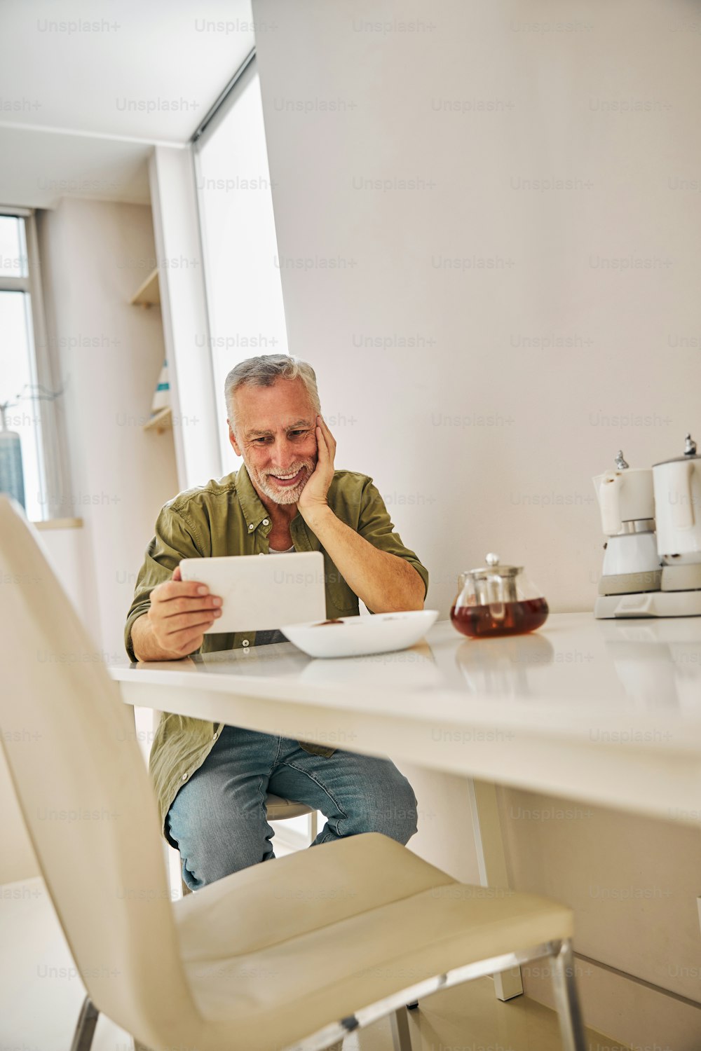 Hombre de edad alegre sonriendo mientras sostiene una tableta y se sienta en la mesa de la cocina con una tetera en ella