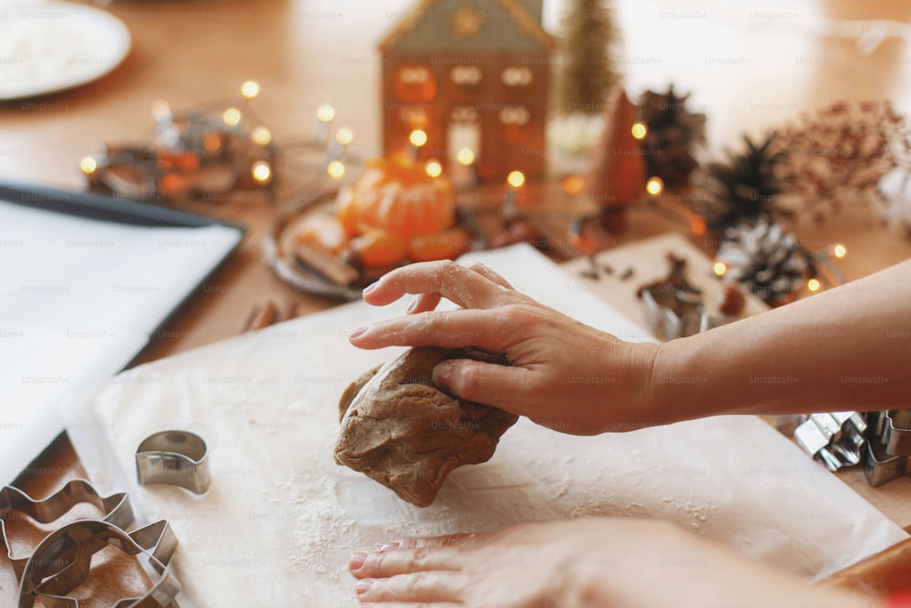 Mãos amassando massa de gengibre cru no fundo de cortadores de metal, especiarias, laranjas, decorações festivas na mesa rústica. Pessoa que faz biscoitos de gengibre, tradição de feriado de Natal