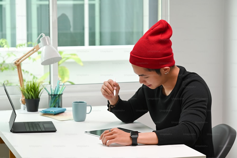 A young male designer in red wool hat working desk with digital graphic tablet and stylus pen at office.