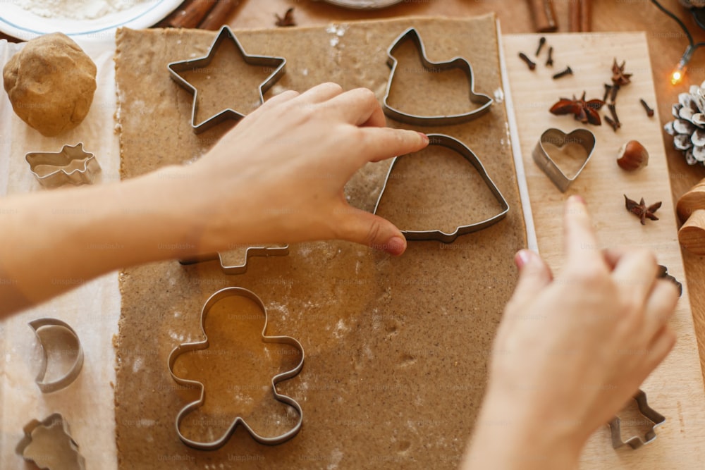 Hands cutting gingerbread dough with festive metal cutters on rustic table with spices, oranges, festive decorations, lights. Person making Christmas gingerbread cookies, holiday advent