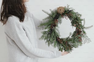 Frau im stylischen Pullover mit modernem rustikalem Weihnachtskranz auf weißem Hintergrund. Rustikaler Weihnachtskranz in weiblichen Händen mit Tannenzweigen, Beeren und Zapfen. Frohe Weihnachten