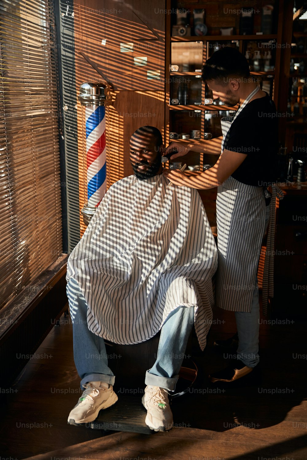 Homem afro-americano barbudo sentado à janela com persianas e sorrindo enquanto o barbeiro retirava a barba de sua bochecha com aparador elétrico