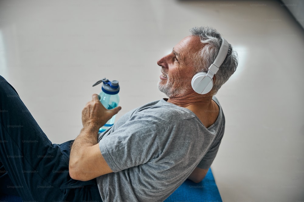 Cropped photo of a cheerful aged man wearing headphones while holding a water bottle and laying on a sports mat