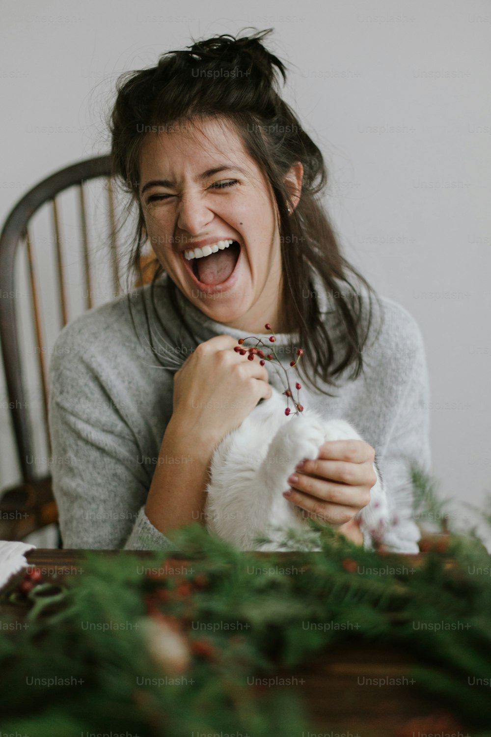 Cute cat helping young happy woman making rustic christmas wreath, holding red berries and green branches. Funny authentic home moments, pet and holidays. Woman laughing with little helper