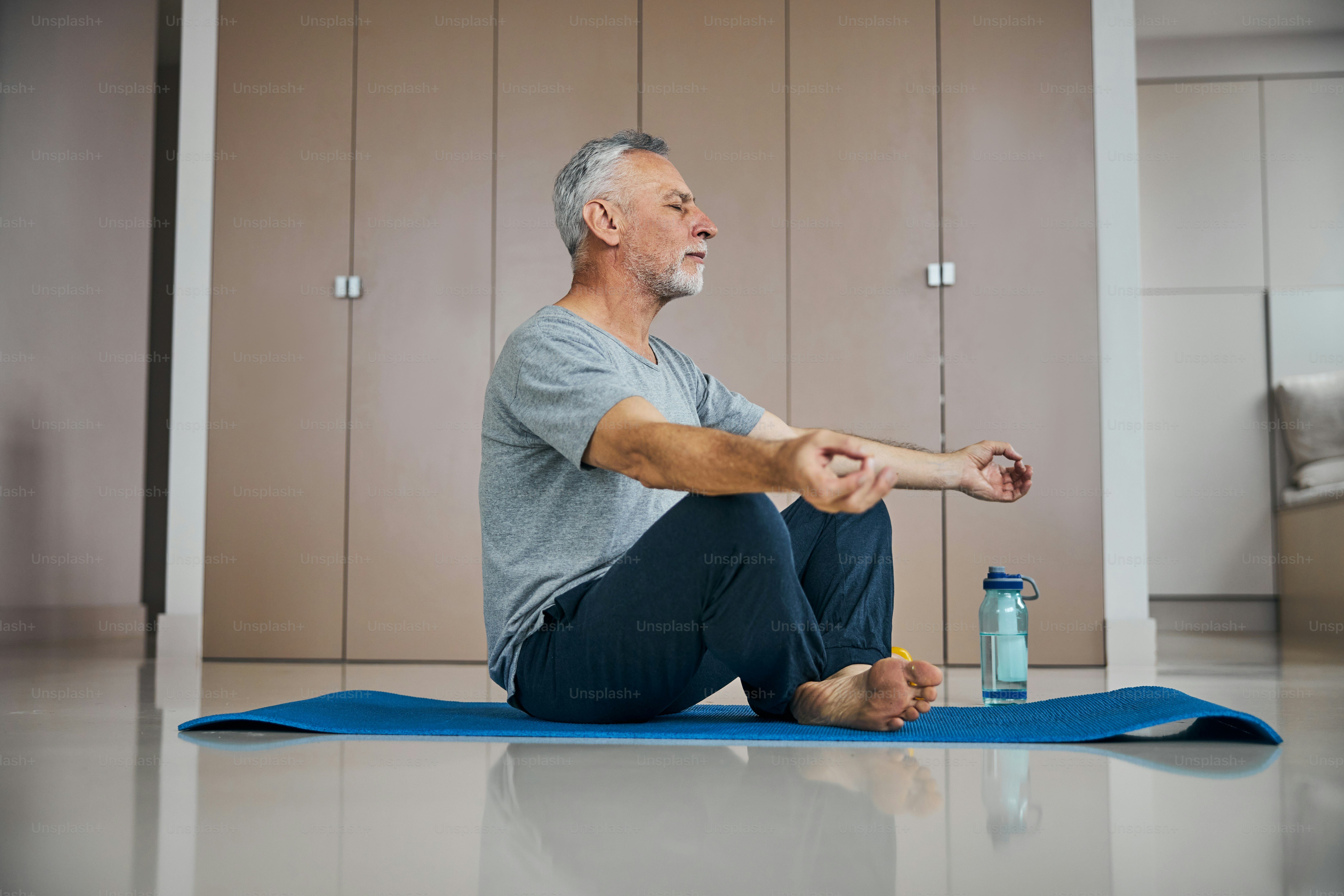 Focused elderly man with his eyes closed sitting on a workout mat while meditating in his living room