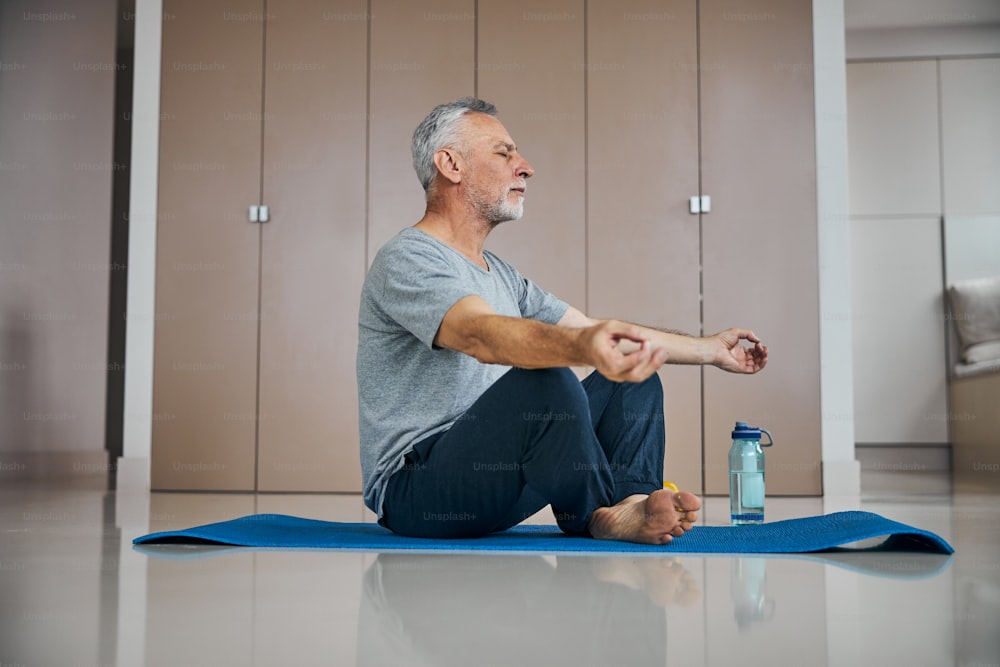 Focused elderly man with his eyes closed sitting on a workout mat while meditating in his living room