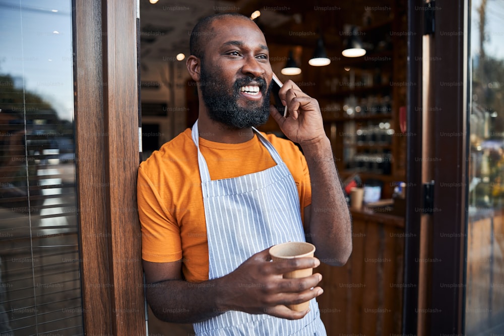 Handsome male worker in apron holding cup of coffee and smiling while having conversation and standing in doorway of barbershop