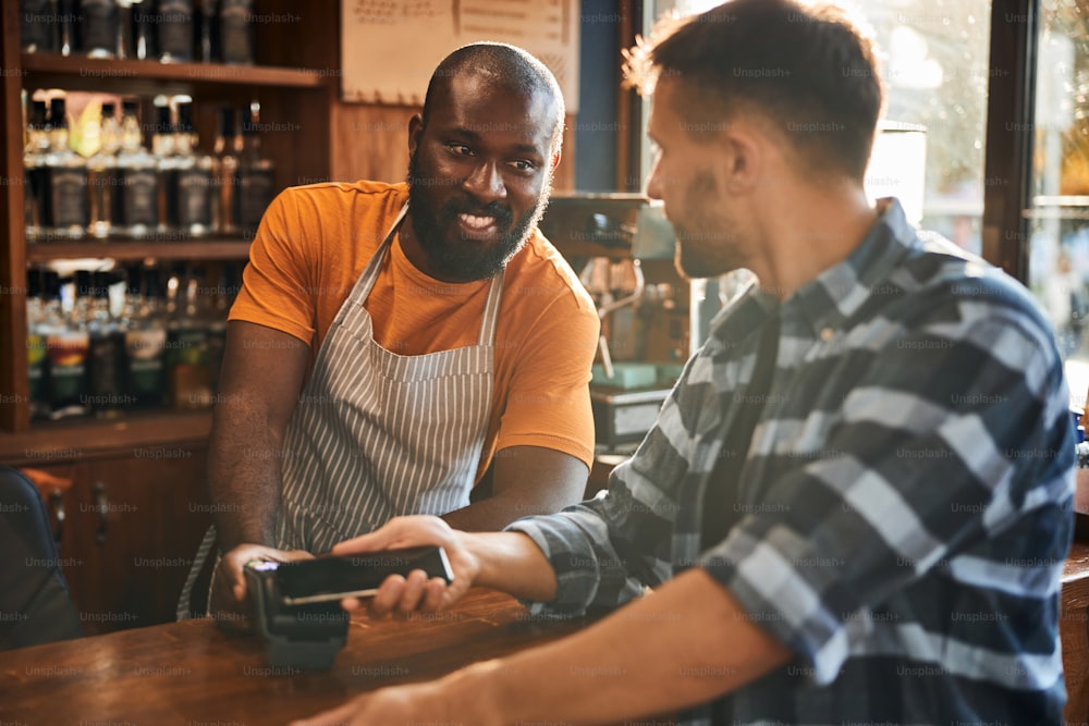 Handsome Afro American man in apron accepting payment from customer while man using smartphone