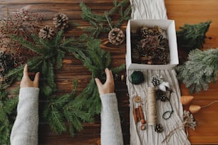 Fazendo rústica coroa de Natal flat lay. Mãos segurando ramos de abeto verde em mesa de madeira rústica com pinhas, bagas, decorações festivas naturais. Workshop sazonal de inverno, advento de férias